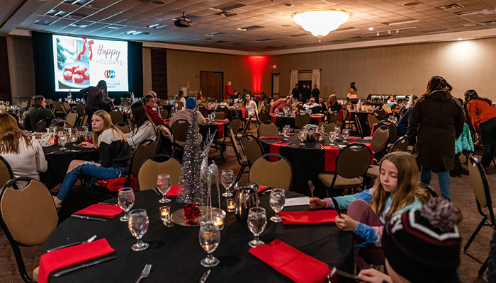 People sit at decorated tables in Rochester Event Center’s banquet hall with a Happy Holidays sign.