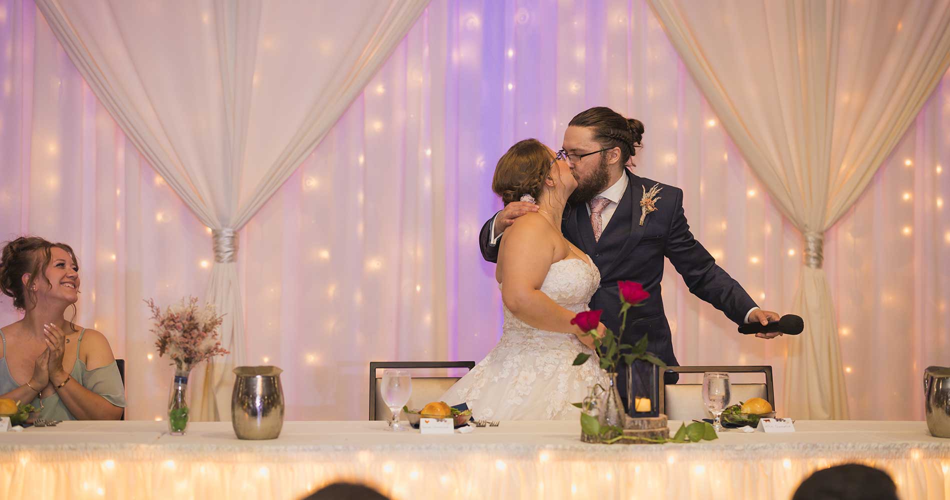 A bride and groom kiss at their wedding reception as a bridesmaid applauds from the table.