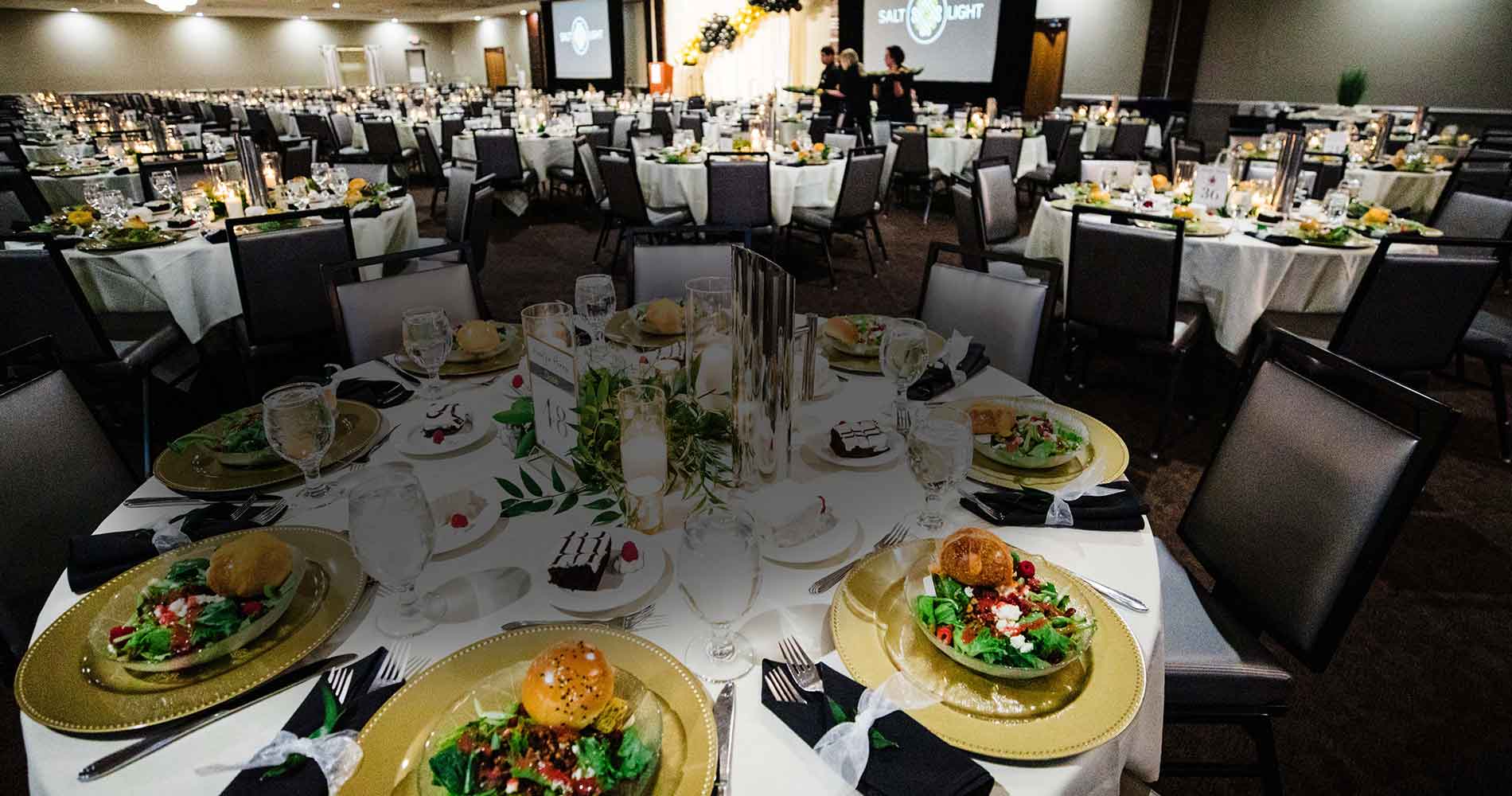 Large banquet hall with round tables set for a formal dinner, salads and rolls on each plate.